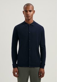 Námořnický modrý svetřík s dlouhým rukávem, pletená textura, zapínání na knoflíky, plochý límec, nošený přes šedé kalhoty. Jednoduchý design s přiléhavým střihem.