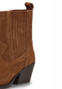 Botín de ante marrón hasta el tobillo con punta afilada, tacón apilado y paneles laterales elásticos. Presenta detalles de costura y una lengüeta en la parte superior.