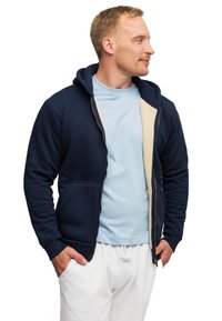 Felpa con zip di colore blu navy, con un morbido rivestimento interno beige, caratterizzata da una tasca frontale e un cappuccio regolabile, indossata sopra una maglietta azzurra chiara.
