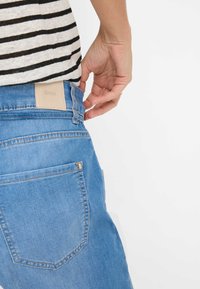 Jean en denim bleu clair avec poches avant et arrière, présentant une étiquette en cuir beige portant la mention "Brax" sur la taille. Haut rayé visible.