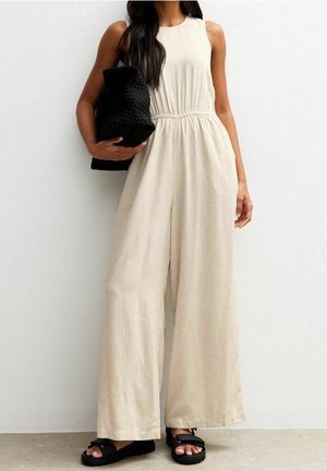 Femme portant une combinaison beige sans manches à jambes larges, tenant une grande pochette noire tissée, accompagnée de sandales noires.