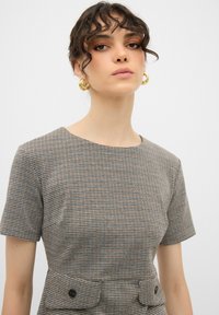 Ruskea ja kerma valkoisella ruudullinen mekko, jossa on lyhyet hihat ja pyöreä kaula-aukko. Mekossa on muotoiltu yläosa ja vyötärövyö mustilla nappilla.