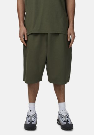 Uomo che indossa pantaloncini larghi verde oliva, top abbinato, calzini bianchi e scarpe da ginnastica grigie e nere, in piedi contro uno sfondo semplice.