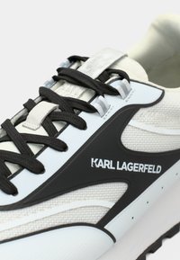 Sapatilha branca e preta com materiais em rede e borracha, com um design curvo, atacadores pretos e a marca "KARL LAGERFELD" na lateral.
