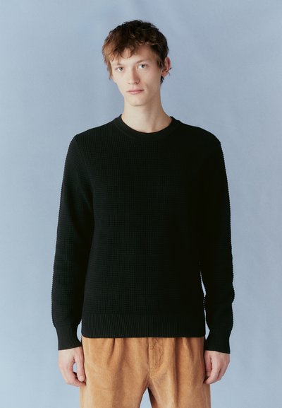 Pulover negru din tricot cu textură, cu guler rotund și manșete cu model în relief, asortat cu pantaloni din corduroy de culoare bej deschis. Fundalul este de culoare albastru deschis.