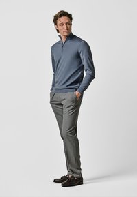 Pull-over zippé gris en tissu doux, associé à un pantalon tailleur gris clair. Le mannequin porte des mocassins en cuir foncé, les mains dans les poches, sur un fond neutre.