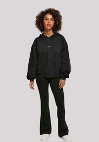 Black hoodie made from soft material, featuring a front pocket, drawstring, and ribbed cuffs. Paired with black flared pants and white sneakers.