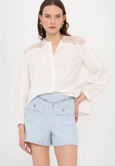 Blusa blanca con detalles de encaje en los hombros, combinada con pantalones cortos de tweed azul claro con dobladillo con flecos y acentos de botón plateado.