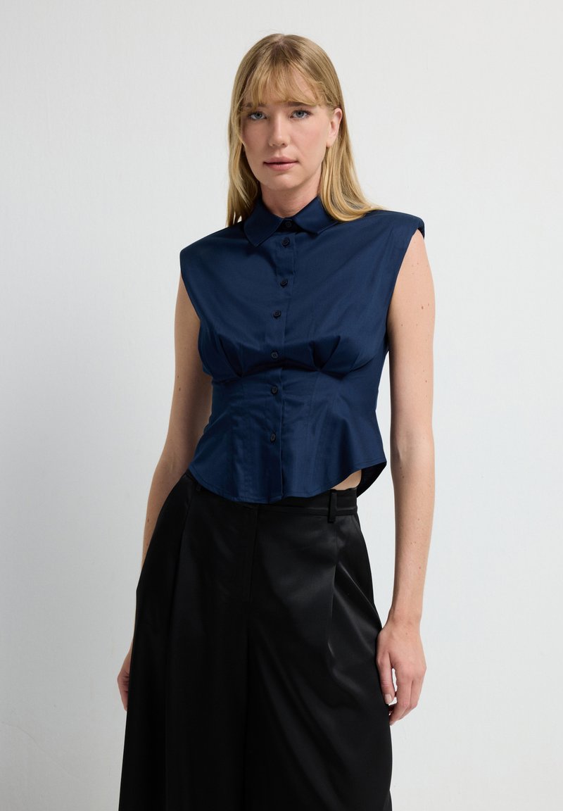 Blusa sin mangas de color azul marino con cuello, cierre de botones y parte delantera con pliegues, combinada con pantalones negros de tiro alto. Textura de tela suave.