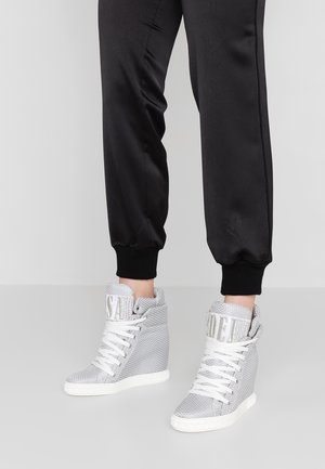 Person iført sorte joggebukser og hvide højtstående kile-sneakers med hvide snørebånd og polstrede kraver.