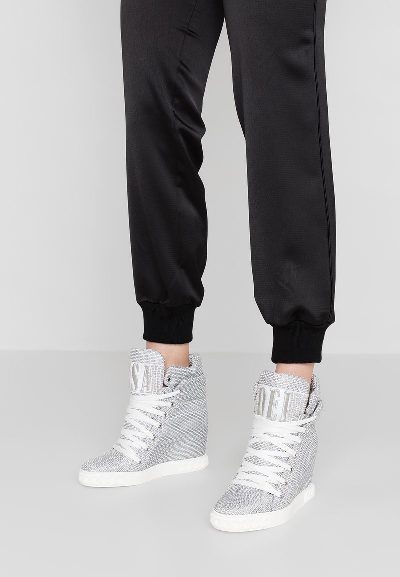 Personne portant un pantalon de jogging noir et des baskets compensées montantes blanches avec des lacets blancs et des cols rembourrés.