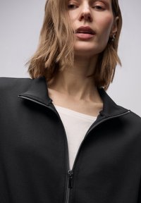 Giacca nera con cerniera, collo alto e tessuto morbido. Indossata sopra una maglia bianca, presenta un design elegante e un dettaglio di texture sottile.