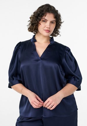 Žena s kučeravými vlasmi nosi tmavomodrú blúzku s volánovým golierom a nadýchanými rukávmi, pózuje na bielom pozadí.