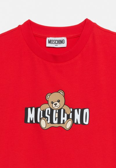 Red kids' T-shirt with ribbed collar featuring a teddy bear graphic holding the white "MOSCHINO" brand name across the chest.