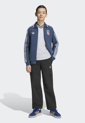 Jonge persoon staand, dragend een blauwe Italië trainingsjas, wit shirt, zwarte broek en blauwe sneakers tegen een effen lichte achtergrond.