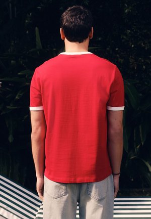 Homme aux cheveux courts et foncés, portant un t-shirt rouge avec un bord blanc et un pantalon gris clair, vu de dos sur un fond sombre avec des feuilles.