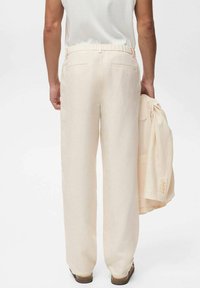Beige Leinenhose mit elastischem Bund und zwei Gesäßtaschen, kombiniert mit einer passenden hellbeigen Jacke. Die Füße sind in braunen Sandalen.