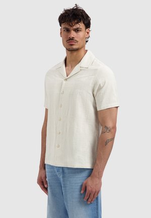 Mann mit lockigem Haar, der ein beige kurzärmeliges Hemd mit dezentem Muster und hellblaue Jeans trägt, steht vor einem schlichten Hintergrund.