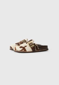 Kuhfell-Muster-Sandalen mit braunen Lederakzenten, ausgestattet mit einem Riemen mit goldfarbenen Beschlägen und flacher Sohle. Texturierte, fellartige Oberfläche.
