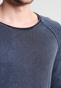 Mörkblå stickad tröja med strukturerad väv, rund halsringning och synliga sömnadsdetaljer på axlarna och ärmarna.