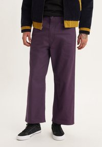 Lila vida byxorna i bomull med en slät textur, med framknäppning och sidfickor, stylade med svarta sneakers.