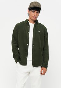 Donkergroene corduroy overhemdd met kraag, gedragen over een wit T-shirt; met witte knopen en een klein logo op de borst.