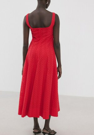 Mujer de pie de espaldas, vistiendo un vestido midi rojo sin mangas con patrón de ojales y sandalias negras de punta abierta sobre un fondo blanco.