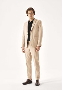 Bež oblečeno obleko s tanko prileganje, enojno gumbnico na blazerju in ujemajoče hlače. Pod njo črna polo majica in rjavi usnjeni čevlji.