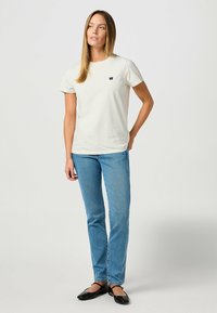 Camiseta de algodón color crema con mangas cortas, cuello redondo y un pequeño logo azul marino en el pecho, combinada con vaqueros azul claro y bailarinas negras.