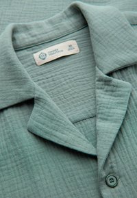 Gros plan sur le col d'une chemise en tissu texturé vert clair avec un bouton et une étiquette indiquant "OMBRE HERITAGE" taille moyenne.