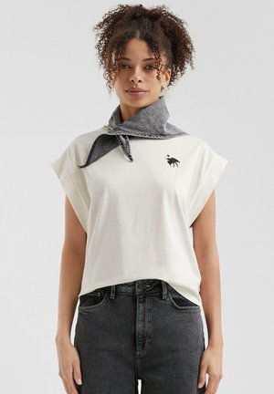 Cremefarbiges Baumwoll-T-Shirt mit kurzen Ärmeln und lockerer Passform, ausgestattet mit einem schwarzen Tiermotiv. Kombiniert mit einem grauen Denimschal, der um den Hals gebunden ist.
