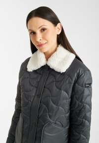 Gequilted zwarte jas met een witte, pluizige kraag en knopen, met zijzakken en een gestikte golfpatroon.
