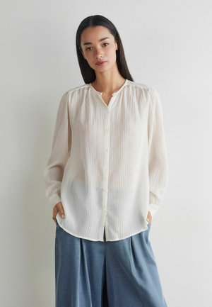 Młoda kobieta z długimi ciemnymi włosami, ubrana w przezroczystą białą bluzkę zapinaną na guziki i luźne niebieskie spodnie, stojąca przy prostej białej ścianie.