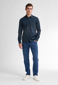 Camicia blu scuro a maniche lunghe con piccoli pois bianchi; presenta colletto button-down e taschino sul petto. Abbinata a jeans azzurri e sneakers bianche.
