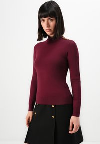 Femme avec des cheveux noirs raides portant un pull col roulé ajusté bordeaux avec des boutons dorés et une jupe noire avec des boutons dorés.