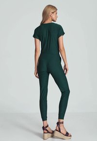 Groene jumpsuit met korte mouwen, taps toelopende benen en zijzakken. Gecombineerd met bruine sleehakken met enkelbandjes en gestructureerde zolen.