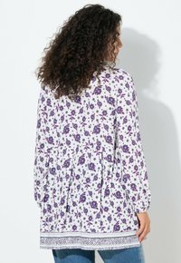 Tunica bianca con motivi floreali viola e blu, maniche lunghe, vita arricciata e bordo decorativo sull'orlo. Tessuto morbido e leggero.