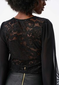 Czarna bluzka z koronką, z przezroczystym, długim rękawem, zdobiona kwiatowymi wzorami i dopasowanym fasonem. Tył prezentuje misternie wykonaną koronkową detalizację.