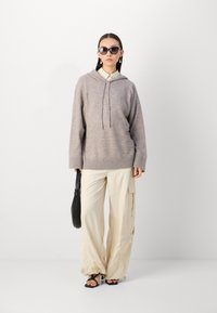 Sudadera de punto marrón claro con cordón, combinada con pantalones cargo beig con bolsillos. Un bolso negro y gafas de sol oscuras completan el conjunto.