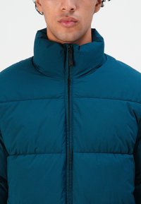 Hombre vistiendo una chaqueta acolchada color aguamarina con un cuello alto y cremallera negra frontal, recortada por debajo de los ojos sobre un fondo blanco liso.