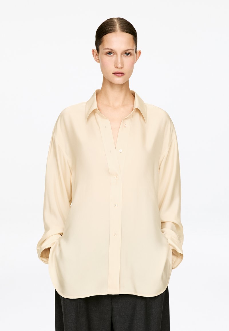 Jeune femme aux cheveux tirés en arrière, portant une chemise oversize couleur crème boutonnée et un pantalon sombre, debout les mains dans les poches.