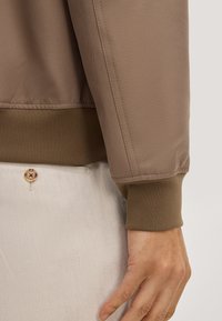Giacca bomber marrone con tessuto strutturato, polsini a coste e finitura liscia. Accenti di filato evidenziano il design. Dettaglio con bottone visibile sui pantaloni.