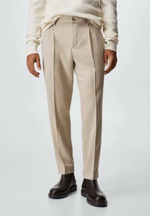 Beige Plissee-Hosen mit taillierter Passform, aus glattem Stoff gefertigt. Verfügen über einen Knopfverschluss und schmal zulaufende Hosenbeine; getragen mit braunen Stiefeletten.