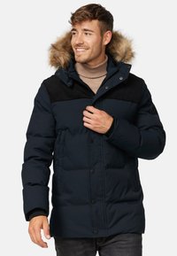 Marineblå varmejakke med et sort fløjlspanel på skuldrene og en hætte foret med faux fur. Har trykknapper og sidelommer.