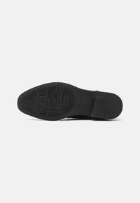 Semelle en caoutchouc noir avec un motif texturé et des bords striés. Présente une forme incurvée et un élément de design central pour une meilleure adhérence.