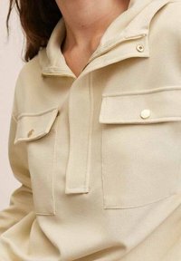 Beiger Pullover mit Reißverschlusskragen, zwei Fronttaschen und goldfarbenen Knöpfen. Strukturierter Stoff mit lässiger Passform und Stichdetails.