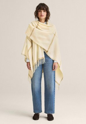 Nainen, jolla on laineikas ruskea tukka ja joka on pukeutunut kermaiseen hapsulliseen huiviin, vaaleansiniseen paitaan, suoralahkeisiin sinisiin farkkuihin ja tummiin mokkasiinikengiin, seisoo paikallaan.