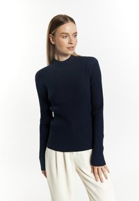 Granatowy sweter z prążkowanego materiału, o wysokim kołnierzu i dopasowanym fasonie. Posiada długie rękawy i lekko rozszerzany dół. W połączeniu z kremowymi spodniami.