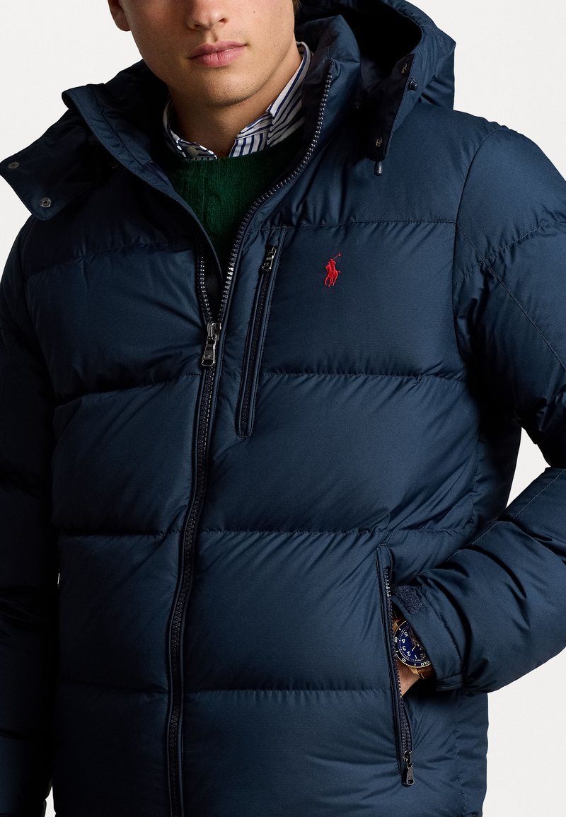 Marineblaue Daunenjacke mit Steppdesign, Reißverschlusstaschen und Kapuze. Sie hat ein rotes gesticktes Logo auf der Brust und eine isolierte Textur.