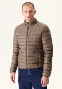 Giacca imbottita marrone con collo alto, zip completa e polsini a coste. Presenta un logo sul braccio sinistro. Indossata sopra jeans blu.